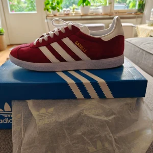 Adidas Gazelle röd/vit mocka sneakers - Säljer ett par Adidas Gazelle sneakers i röd mocka med vita detaljer och klassiska tre ränder på sidorna. Skorna har vit sula, snörning och guldfärgad Gazelle-text på sidan. Perfekta för dig som vill ha en ikonisk och stilren sneaker med retrovibbar.
