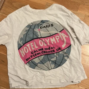 Vit t-shirt Uniqlo x Olympia Le-Tan - Vit t-shirt från Uniqlo i samarbete med Olympia Le-Tan. Stor tryckt grafik på framsidan med texten 'Hotel Olympia Paris' i rosa och grått. Klassisk passform med rund hals och korta ärmar. Skön bomullskvalitet, perfekt för en chill look.