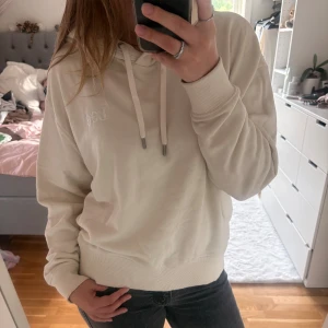 Beige hoodie från Lee med huva - Säljer en beige hoodie från Lee med broderad logga på bröstet. Tröjan har huva med snörning, ribbade muddar och en avslappnad passform. Perfekt för mysiga dagar och enkel att matcha med jeans eller leggings.