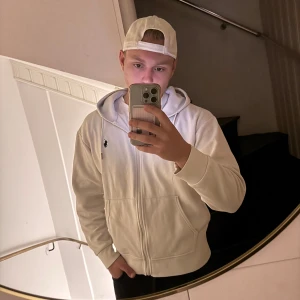 Vit hoodie från Ralph Lauren - Vit hoodie från Ralph Lauren med dragkedja och snörning i huvan. Klassisk svart logga på bröstet och två fickor framtill. Skön passform och perfekt för chill dagar. Materialet är mjuk bomull och hoodien har långa ärmar. En synlig fläck andra fläckarna på bröstet märks knappt samt smink stänket på armen, pris kan diskuteras 