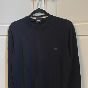 Blå stickad tröja från BOSS - Blå stickad tröja från BOSS med diskret logga broderad på bröstet. Tröjan har rundad hals med ribbad kant och långa ärmar. Perfekt för dig som gillar stilrena och klassiska plagg med snygg passform.