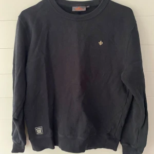 Svart sweatshirt från Morris, M - Svart sweatshirt från Morris med rund halsringning och broderad logga på bröstet. Tröjan har ribbade muddar vid ärmslut och nederkant, samt en liten patch nertill. Perfekt för en clean och stilren look.