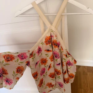 Blommig offshoulder topp från H&M Divided - Superfin topp från H&M Divided med blommigt mönster i orange och rosa på en ljusbeige botten. Toppen har vida ärmar, smala axelband som korsas i ryggen och är gjord i ett lätt, luftigt material. Perfekt för sommarens alla tillfällen!