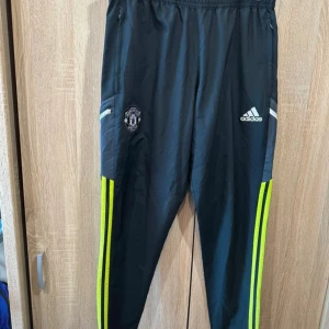 Manchester United Adidas träningsbyxor M - Svarta träningsbyxor från Adidas med Manchester United-logga på benet och neongula ränder längs sidorna. Byxorna har elastisk midja, dragkedjor vid fickorna och reflekterande detaljer baktill. Perfekta för fotboll eller träning.