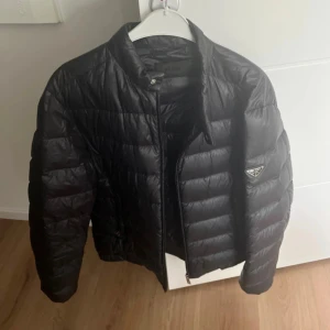 Svart pufferjacka från Prada - Säljer en svart pufferjacka från Prada med klassisk quiltad design och diskret logga på ärmen. Jackan har dragkedja framtill, ståkrage och tryckknappar vid fickorna. Perfekt för dig som vill ha en stilren och lyxig look.