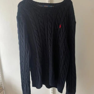 Marinblå kabelstickad tröja Polo Ralph Lauren - Kabelstickad svart tröja från Polo Ralph Lauren med klassisk rund hals och röd broderad logga på bröstet. Långärmad och i mjuk bomull, perfekt för lager-på-lager under höst och vinter. Snygg och tidlös modell som funkar till allt. Köpte den för cirka 2200 kr ny två veckor sedan på nätet och märkte att den var för stor. Den är använd 1 gång 