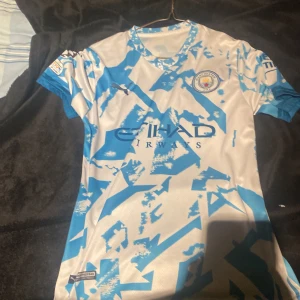 Manchester City De Bruyne fotbollströja - Säljer en Manchester City fotbollströja från Puma med De Bruyne och nummer 17 på ryggen. Tröjan är vit med blått abstrakt mönster, kortärmad och har officiella UEFA Foundation och Puma-loggor. Materialet är lätt och andas, perfekt för match eller träning.