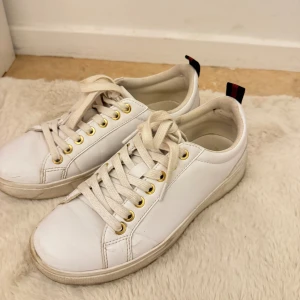 Vita sneakers med gulddetaljer - Snygga vita sneakers i skinn med guldfärgade öljetter och vita snören. Baktill finns en svart och röd randig detalj med guldfärgad knapp. Klassisk rund tå och platt sula, perfekt för en clean och stilren look.