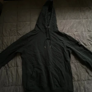 Mörk grön hoodie från Calvin Klein Jeans - Svart hoodie från Calvin Klein Jeans med huva och dragsko. Klassisk känguruficka framtill och snygg CK-logga på vänster ärm. Mjuk bomullskvalitet och ribbade muddar vid ärmslut och nederkant. 