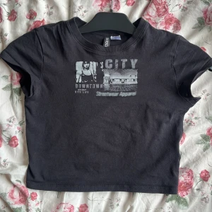 Svart croppad t-shirt med citytryck, baby tee  - Svart croppad t-shirt från Bik Bok med grått cityinspirerat tryck framtill. Toppen har rund halsringning och korta ärmar, perfekt för en urban och avslappnad stil.