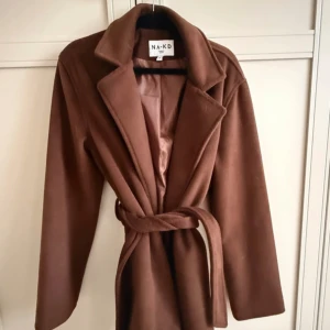 Brun kappa med bälte från NA-KD - Snygg brun kappa från NA-KD med klassisk krage och bälte i midjan. Jackan har lång ärm och en stilren, enkel design som passar perfekt till höst och vinter. Materialet är mjukt och ger en lyxig känsla. Perfekt för dig som gillar tidlös stil.