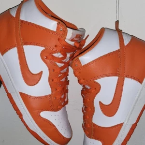 Nike Dunk High Orange/White - Snygga Nike Dunk High  Syracuse. Perfekt för att komplettera din sneaker-kollektion eller för att ge din outfit en färgstark touch. Använda men mycket bra skick där av priset Storlek.42 Kommer utan box Kolla gärna mina andra annonser