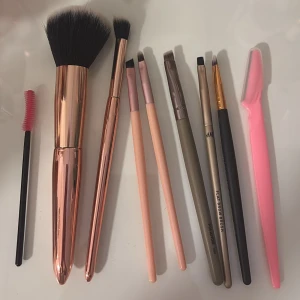Set med sminkborstar och ögonverktyg - Säljer ett kit med sminkborstar i roséguld och pastellfärger, inklusive stor puderborste, mindre borstar för ögonskugga och bryn, samt en rosa ögonfransborste och en rosa ansiktsrakkniv. Perfekt för att fixa både bas och detaljer.