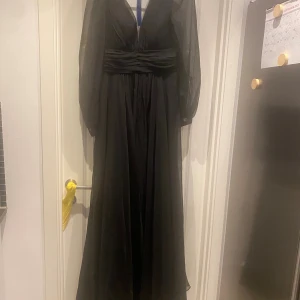 Svart långklänning från Cinderella Divine - Elegant svart långklänning från Cinderella Divine med v-ringning, markerad midja och långa transparenta ärmar. Klänningen har ett svepande fall och är tillverkad i ett tunt, lätt material som ger en lyxig känsla. Perfekt för bal eller festliga tillfällen.