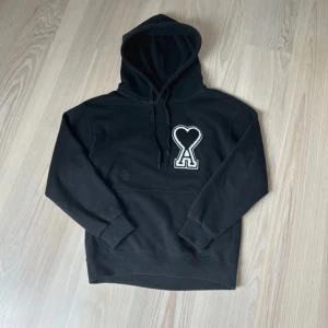 Svart hoodie från Ami med hjärtdetalj - Svart hoodie från Ami med stor vit och svart hjärtdetalj och bokstaven A på bröstet. Tröjan har huva med snörning, känguruficka och ribbade muddar. Tillverkad i mjuk bomull, perfekt för chill dagar. Snygg och stilren design som sticker ut.