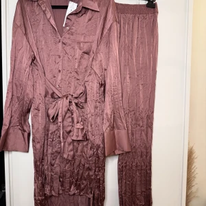 Mauve pyjamasset från Gina Tricot M - Snyggt tvådelat set från Gina Tricot i färgen Wistful Mauve. Setet består av en långärmad skjorta med knappar, krage och knytband i midjan samt matchande vida byxor med resår i midjan. 