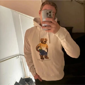 Vit hoodie Polo Ralph Lauren björn - Vit hoodie från Polo Ralph Lauren med ikoniskt björntryck på bröstet. Tröjan har huva med snörning och långa ärmar. Tillverkad i mjuk bomull, perfekt för en chill och stilren look. Små fläckar på ena armen