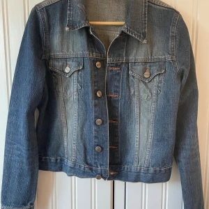 Jeansjacka - Snygg blå jeansjacka med klassisk passform och silvriga knappar framtill. Jackan har två bröstfickor med lock och knapp, samt långa ärmar.