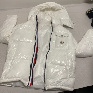 Vit pufferjacka från Moncler - Snygg vit pufferjacka från Moncler med glansig finish, huva och dragkedja framtill. Jackan har en bröstficka med Moncler-logga och coolt innerfoder med serietidningsmotiv. Röd, vit och blå detalj längs dragkedjan ger extra stilpoäng.