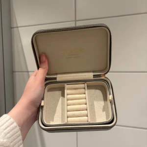 Sif Jakobs smyckesväska i silver - Stilren smyckesväska från Sif Jakobs i metallic silver med dragkedja runtom. Insidan är ljusbeige och har fack för ringar och smycken. Perfekt att ha med på resan eller för att hålla ordning på dina favoritsmycken.