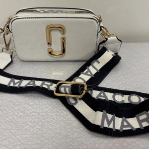 Vit axelväska från Marc Jacobs - Snygg vit axelväska från Marc Jacobs med guldiga detaljer och dubbla dragkedjor. Väskan har en bred, svartblå och vit axelrem med logotypen tryckt i grått. Rektangulär form och stilren design, perfekt för att lyfta din outfit. I mycket fint skick. Har fått i present, kan ej garantera original. Skickas eller hämtas i Göteborg 