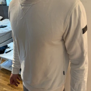 Vit sweatshirt från Strömberg i bomull - Vit långärmad sweatshirt från Strömberg i mjuk bomull. Tröjan har rund halsringning, ribbade muddar och en snygg svart logotyp-patch på ärmen. Enkel och stilren modell som passar perfekt till jeans eller joggers.