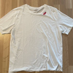Vit t-shirt från Saint Laurent - Vit t-shirt från Saint Laurent med en liten röd broderad läppdetalj vid axeln. Klassisk rund halsringning och korta ärmar. Tillverkad i mjuk bomull för en skön känsla och clean look.