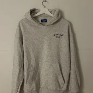 Grå/vit hoodie från Jack & Jones - Snygg grå hoodie från Jack & Jones med trycket 'ATHLETIC CLUB 1990' på bröstet. Tröjan har huva, magficka och långa ärmar. Perfekt för chill dagar och enkel att matcha med jeans eller joggers. Mjuk insida och klassisk passform.