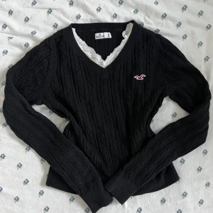 Svart vintage stickad Hollister v-ringad tröja M - Svart vintage kabelstickad tröja från Hollister med v-ringning och lång ärm. Tröjan har ett litet rött Hollister-logo broderat på bröstet och ribbade muddar vid ärmslut och nederkant. Snyggt mönster och klassisk passform, perfekt för lager-på-lager. Spetslinne ingår ej. Måtten finns vid behöv. 