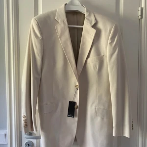 Ecru kavaj från Dressmann Batistini - Snygg ecrufärgad kavaj från Dressmann Batistini i bomull. Klassisk modell med två knappar framtill, fyra ljusa knappar vid ärmslut och två fickor med lock. Perfekt för dig som gillar stilrena och ljusa plagg. Passar till både skjorta och t-shirt.