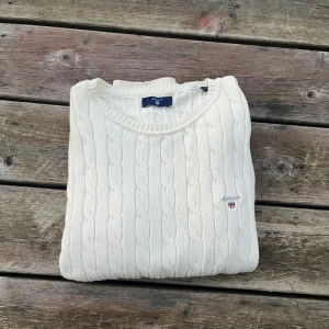 Vit kabelstickad tröja från Gant - Kabelstickad vit tröja från Gant med rund halsringning och broderad logga på bröstet. Tröjan är i mjuk bomull och har ribbade muddar vid ärmslut och nederkant. Perfekt för en clean och klassisk look. Perfekt för hösten 🍁🍂