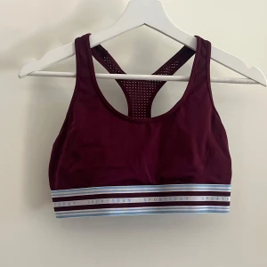 Vinröd sporttopp från H&M Sport - Vinröd badtopp från H&M Sport med brottarrygg och ventilerande meshdetaljer. Elastiskt band med blå, vita och vinröda ränder och texten 'SPORTSDAY' runt om. 