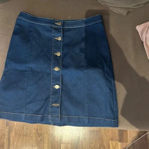 Mörkblå jeanskjol från Jane Skirt - Snygg mörkblå jeanskjol från Jane Skirt i mid rise-modell. Kjolen har raka linjer, dekorativa guldknappar framtill och kontrastsömmar. Perfekt för dig som gillar klassisk denimstil och vill ha en enkel men trendig kjol.
