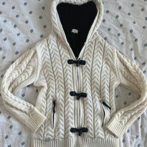 Vit stickad hoodie från Hollister - Vit kabelstickad hoodie från Hollister med mysig svart fleecefodring. Tröjan har huva, dragkedja och snygga knappar framtill samt två fickor. Perfekt för kyliga dagar när du vill vara både varm och stilren. Dragkedjan funkar men saknar delen man dra i. Kan skicka bild vid behöv. 