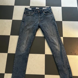 Blå jeans från H&M med rak passform - Säljer ett par klassiska blå jeans från H&M med rak passform och fem fickor. Jeansen har en mellanblå tvätt och traditionell knappgylf. Skinnpatch med H&M-logga baktill i midjan. Perfekta till sneakers eller boots.