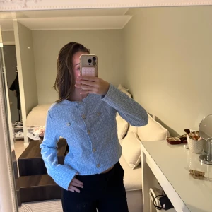 Ljusblå ribbad kofta med knappar - Säljer en ljusblå croppad kofta med ribbad struktur och snygga guldfärgade knappar framtill. Koftan har lång ärm och rund hals, samt dekorativa fickor med knappar på bröstet. Kan även användas som en kofta 