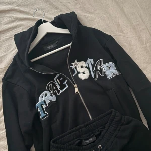 Trapstar dress - Svart hoodie från Trapstar med stor broderad logga i blått, vitt och silver på bröstet. Tröjan har huva, dragkedja framtill och långa ärmar. Perfekt för en streetwear-look och riktigt skön att bära.