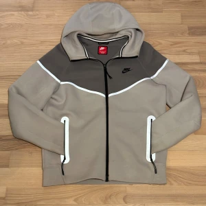 Nike tech fleece reflex  - Nike hoodie i beige och grå med svart logga på bröstet. Tröjan har hel dragkedja, huva och vita detaljer längs bröst och fickor. Tillverkad i mjukt material, perfekt för chill eller träning. Snygg och stilren design med långa ärmar och praktiska fickor.