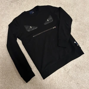 Fendi tröja  - Super cool svart tröja från Fendi med unika detaljer. Tröjan har en dragkedja framtill och coola läderdetaljer som ger ett edgy utseende. Perfekt för dig som vill sticka ut med stil. Storlek S men kan även passa dig som har M. 