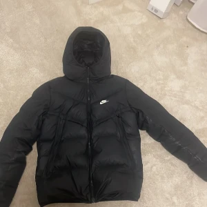 Svart Nike pufferjacka Storm-FIT - Svart pufferjacka från Nike med Storm-FIT-teknologi. Jackan har huva, två snedställda dragkedjefickor framtill och klassisk Nike-logga på bröstet. Materialet är syntet och jackan är vadderad för extra värme. Perfekt för kalla dagar.