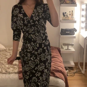 Svart blommig omlottklänning - Svart långklänning med vita blommor och omlottmodell. Klänningen har v-ringning, trekvartsärm och knyt i midjan för en snygg passform. Perfekt för dig som gillar en feminin och trendig stil.