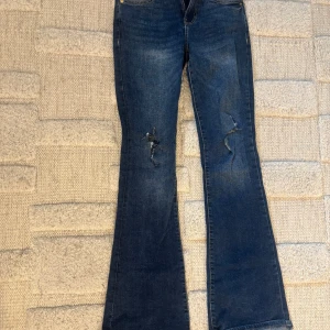 Blå bootcut jeans från Bershka - Snygga mörkblå jeans från Bershka med bootcut passform och slitna detaljer på benen. Klassisk femficksmodell med guldfärgade nitar och knapp. Jeansen har låg/normal midja och är tillverkade i jeansmaterial med lite stretch. Tveka inte på att höra av dig vid frågor eller intresse!