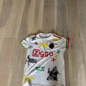 Ajax x Bob Marley vit fotbollströja Adidas - Ajax x Bob Marley special edition fotbollströja från Adidas i vit polyester med färgglada detaljer på ärmarna. Tryck med Ziggo-logga, Ajax-märke och Bob Marley-inspirerade grafiska prints och texter. Kortärmad modell med rund hals och Aeroready-material.