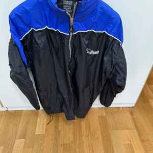 Bear vindjacka blå och svart - Säljer en retro vindjacka från Bear med blått parti upptill och svart nedtill. Jackan har vit piping, dragkedja framtill, hög krage och broderad logga på bröstet och ryggen. Materialet är lätt och syntetiskt, perfekt för blåsiga dagar.