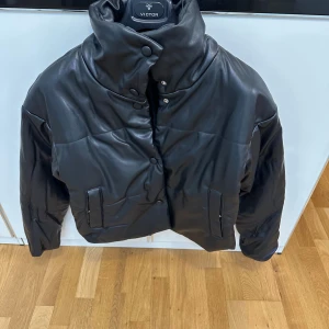 Svart pufferjacka i skinnlook - Säljer en svart pufferjacka med hög krage och snygg skinnlook. Jackan har knappar framtill, två fickor och en riktigt chunky vibe. Perfekt för kyliga dagar