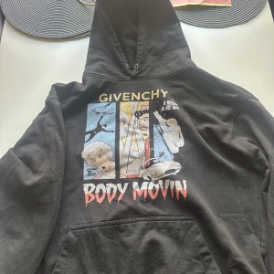 Svart hoodie Body Movin - Svart hoodie med stor, färgglad grafisk print på framsidan och texten 'BODY MOVIN'. Tröjan har huva, känguruficka och långa ärmar. Trycket har retro-inspirerade motiv i blått, gult, rött och vitt. Perfekt för dig som gillar statement-plagg och streetwear.