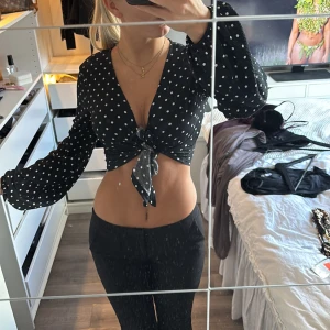 Svart prickig croppad blus med knyt - Säljer en svart croppad blus med vita prickar och v-ringning. Toppen har långa puffärmar med resår vid ärmslut och snygg knytning framtill. Materialet är lätt och luftigt, perfekt för att skapa en trendig look. Passar dig som gillar att sticka ut med coola detaljer. Osäker om jag vill sälja