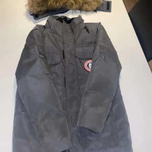 Grå parkajacka Canada Goose - Säljer en grå parkajacka från Canada Goose med avtagbar huva och fuskpäls. Oanvänd, Jackan har flera fickor framtill, dragkedja och kardborreknäppning. Perfekt för kalla dagar och har klassisk patch på bröstet.