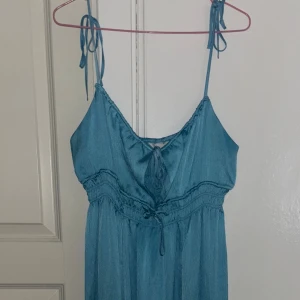 Blå omlottklänning med knytband - Supersöt blå klänning med tunna axelband som knyts på axlarna och omlottdetalj framtill. Klänningen har smockad midja och lätt, flowigt tyg som ger en chill vibe