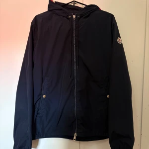 Moncler Windbreaker  - Snygg mörkblå vindjacka från Moncler med huva och dragkedja framtill. Jackan har två fickor med tryckknappar, justerbar dragsko i huvan och Moncler-logga på ärmen. Små defekter förekommer därav det sjukt bra priset!